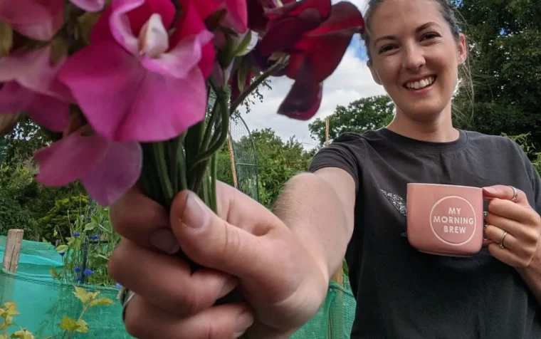 Woman holding vibrant pink sweet peas in a garden, smiling whilst displaying a 'My Morning Brew' mug among flowering plants.