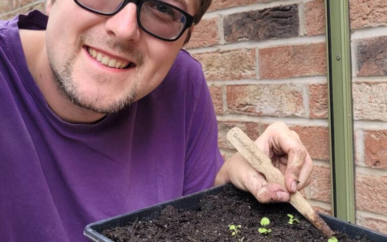 Chris Jesson, PlantGrow gardening champion, proudly holds a tray of seedlings in natural compost, smiling against a brick wall backdrop.